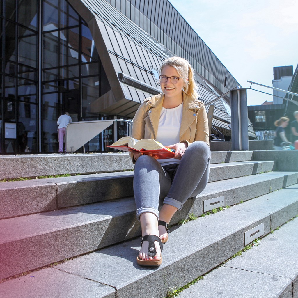 Eine Frau mit blonden Haaren, die zu einem Pferdeschwanz gebunden sind, sitzt vor der Münsteraner Stadtbücherei und liest in einem Buch.