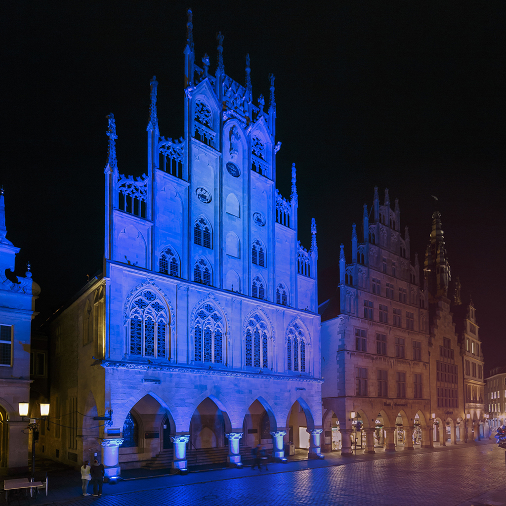 Das blau angeleuchtete Rathaus am Prinzipalmarkt in Münster und die danebenliegenden historischen Fassaden bis zum Stadthausturm.
