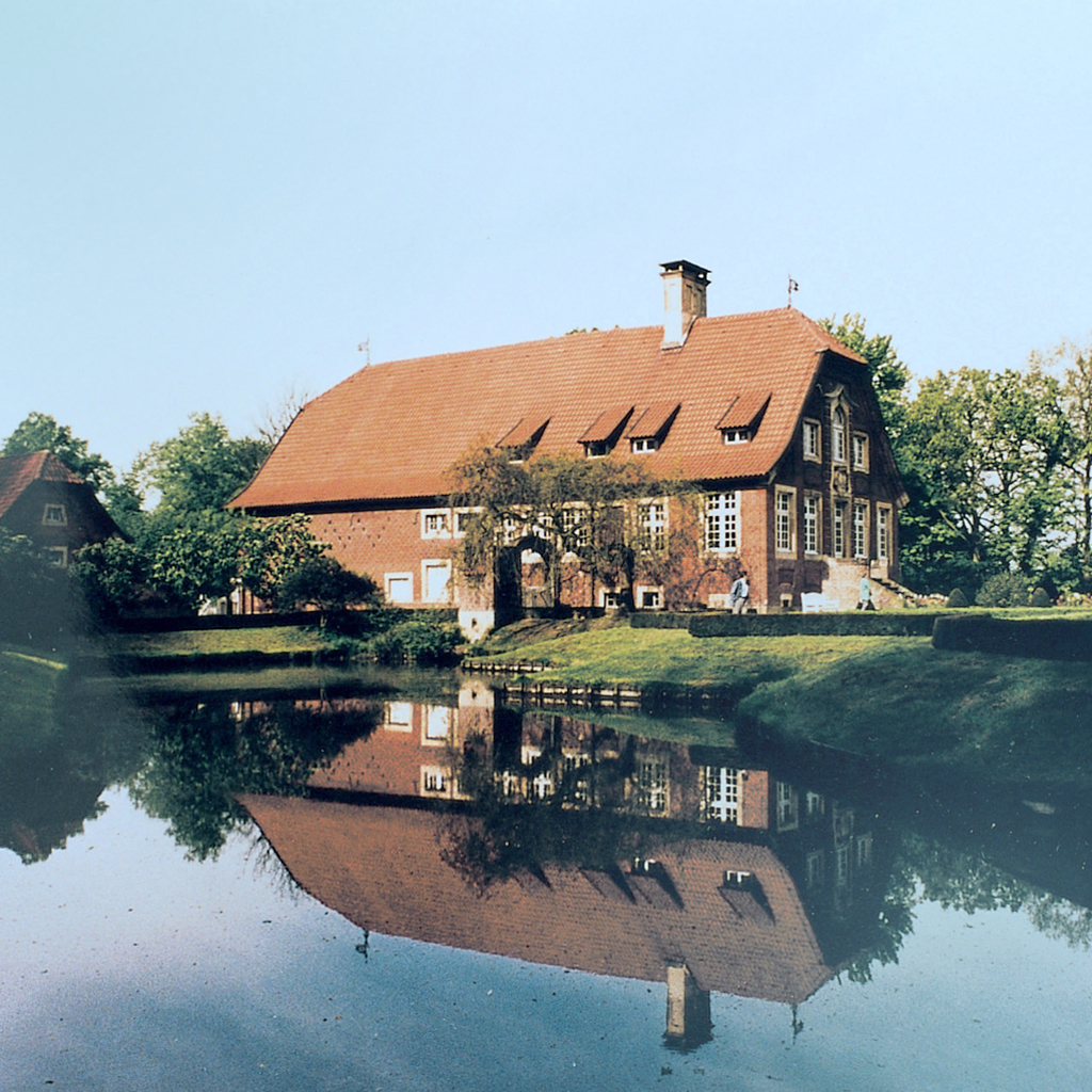 Das abgebildete Haus Rüschhaus war das Wohnhaus der Dichterin Annette von Droste-Hülshoff und spiegelt sich unter blauem Himmel in einem See.