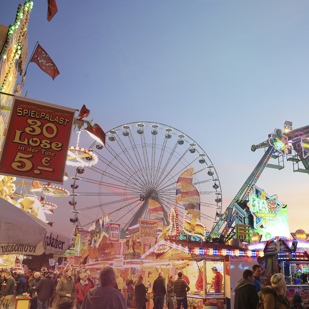 Ein erleuchtetes Riesenrad und zahlreiche Kirmesbuden sowie ein weiteres Fahrgeschäft und eine Losbude im Vordergrund zeigen den Send in Münster.