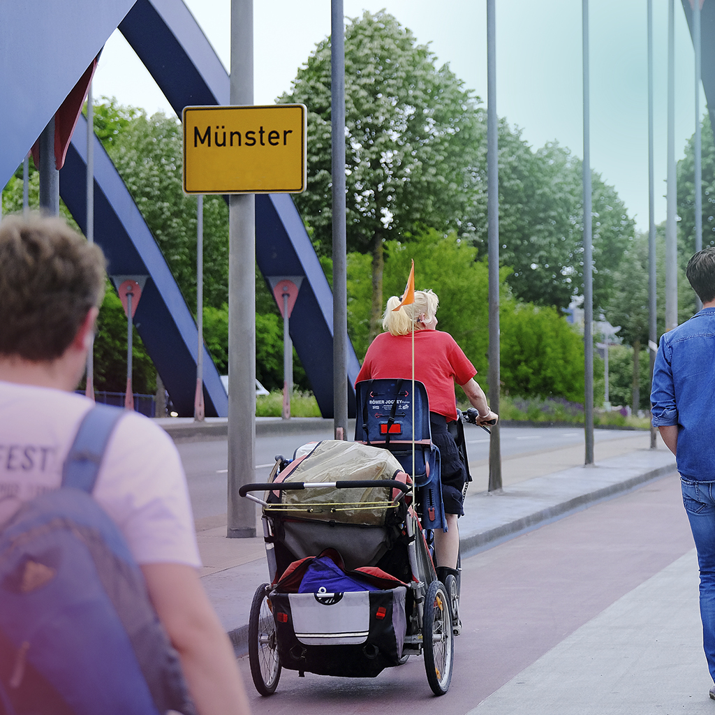 Ein Ortsschild an einer befahrenen Straße schildert Münster aus, im Vordergrund sind verschiedene Menschen zu sehen, die zu Fuß oder mit dem Fahrrad unterwegs sind.