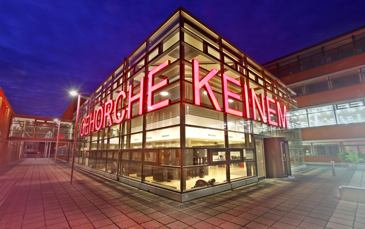 Die Universitätsbibliothek in Münster mit effektvoller Beleuchtung, wobei in hellroten Leuchtbuchstaben "gehorche Keinem" auf der Fassade steht.