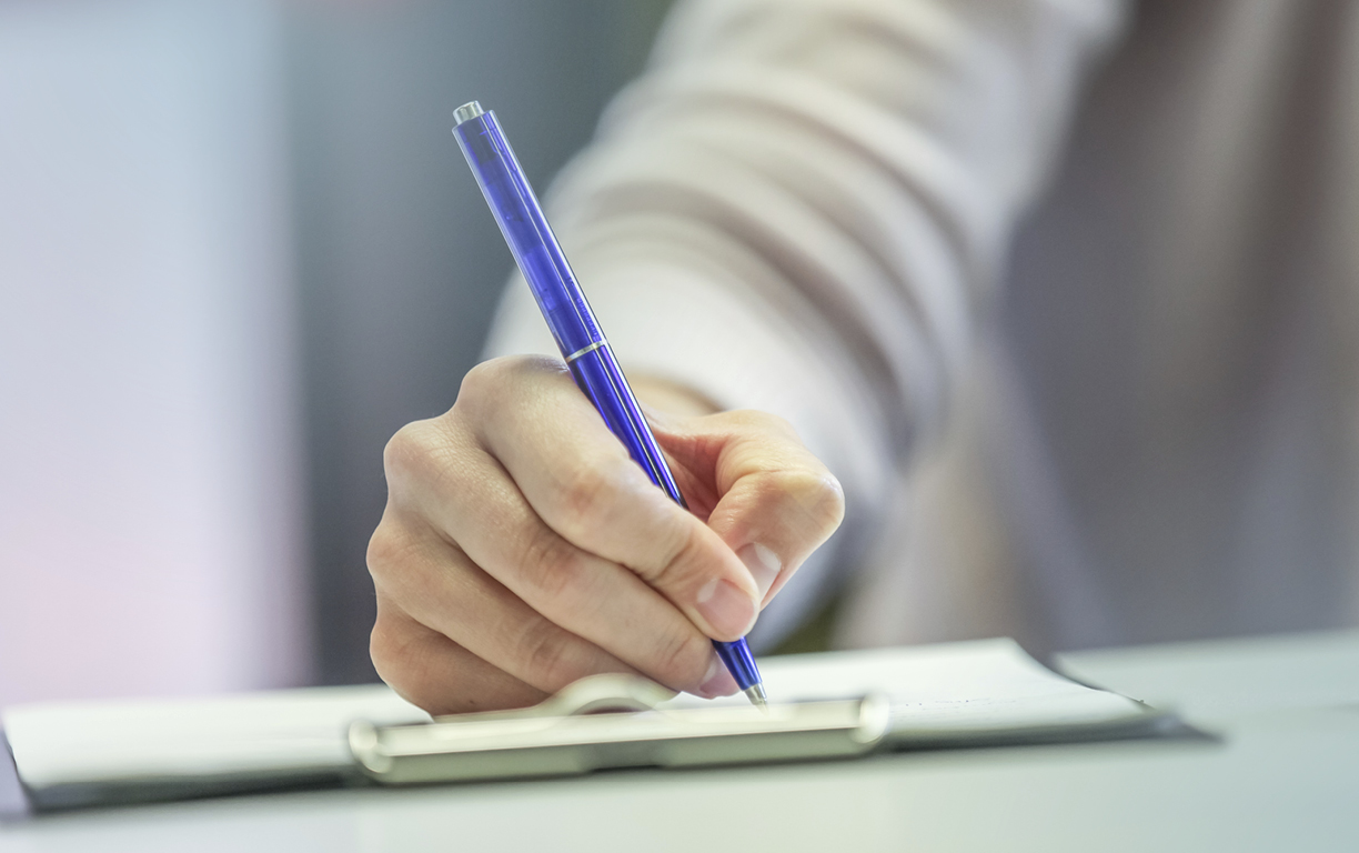 Eine Hand jält einen blauen Kugelschreiber füllt dabei schreibend einen Zettel auf einem Klemmbrett aus.