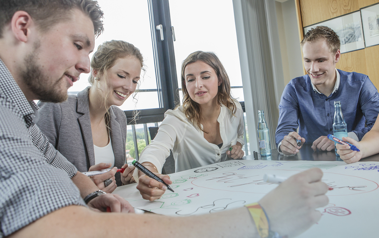 Eine Gruppe mit vier bunt gekleideten jungen Menschen, die gemeinsam schriftlich auf ein vor ihnen liegendes Plakat Ideen sammeln.Sammeln der Erfahrungsberichte