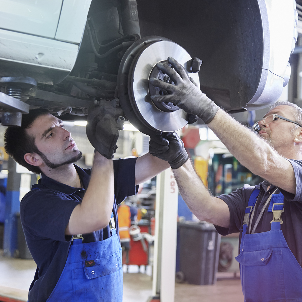 Zwei Industriemechaniker im Blaumann arbeiten an einem Rad eines Fahrzeugs, welches auf einer Hebebühne steht.