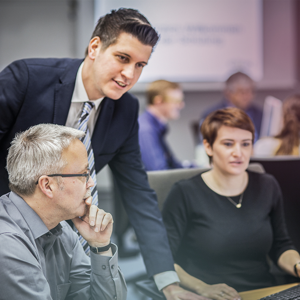 Ein Kaufmann für Digitalisierungsmanagement trägt einen Anzug und berät einen Mann im Vordergrund und eine Frau im Hintergrund, welche beide Anwender/-innen von IT-Systemen sind und auf einen Computer schauen.