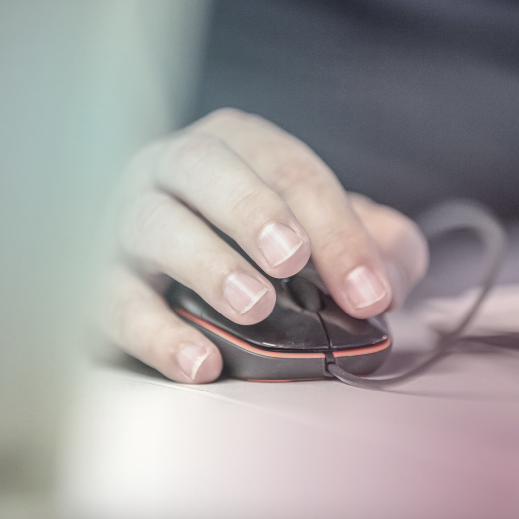 Eine Hand führt eine  schwarze Computermaus, die auf einem weißen Tisch liegt, der Zeigefinger der Hand klickt auf die linke Maustaste.