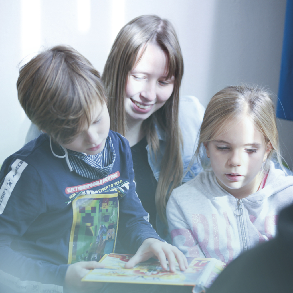 Eine Erzieherin sitzt hinter zwei Kindern und liest einem Jungen und einem Mädchen etwas aus einem Kinderbuch vor.
