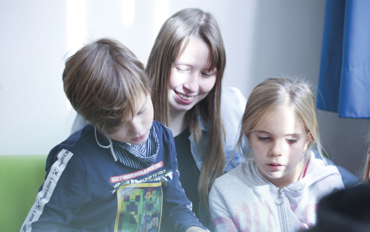 Eine Erzieherin sitzt hinter zwei Kindern und liest einem Jungen und einem Mädchen etwas aus einem Kinderbuch vor.