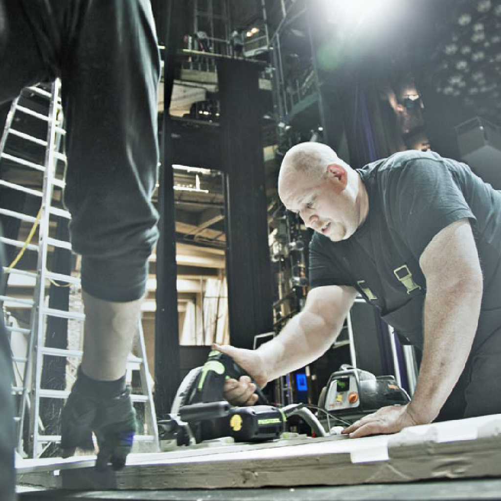 Ein Veranstaltungstechniker baut im Vordergrund ein Bühnenbild im Theater Münster auf, Hände eines Kollegen helfen dabei.
