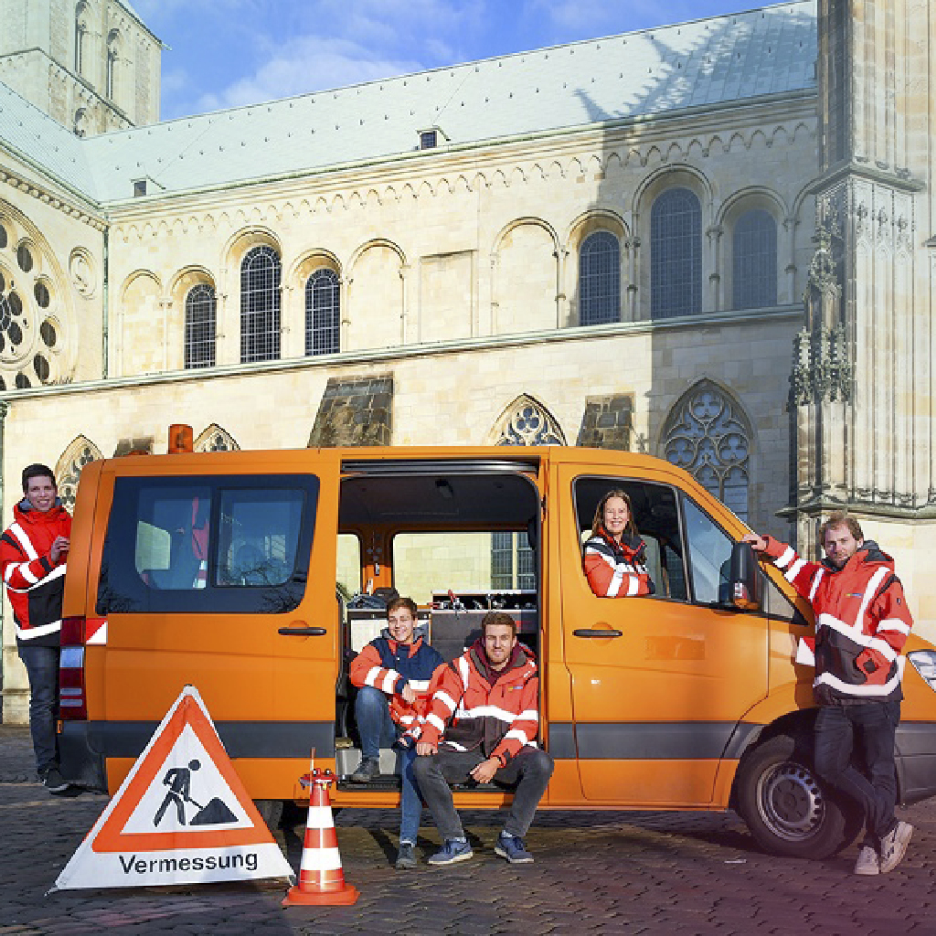 Vor dem St.-Paulus-Dom in Münster steht ein oranges Vermessungsfahrzeug der Stadt, in und um das Fahrzeug herum stehen fünf Arbeiter/-innen mit orangen Westen.