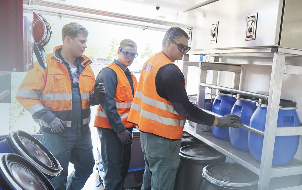 Drei Männer mit orangen Warnwesten stehen in einem Kraftfahrzeug für Schadstoffe und sortieren verschiedene Gefäße und Tonnen.