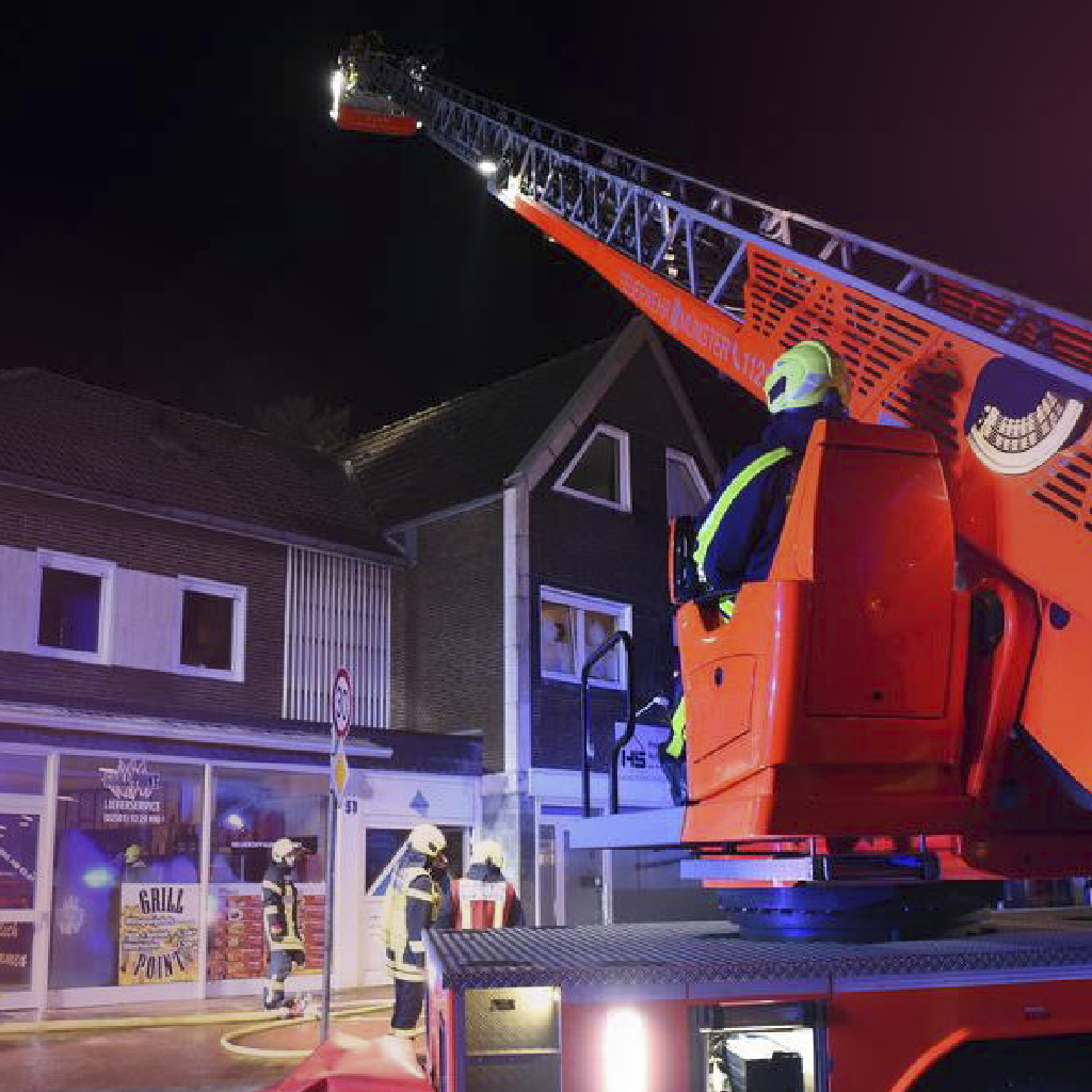 Ein Brandmeister steht auf einem Kran eines Feuerwehrautos, welcher auf ein Wohnhaus gerichtet ist, weitere Feuerwehrmänner und -frauen stehen auf der Straße vor dem Haus und sichern diese.