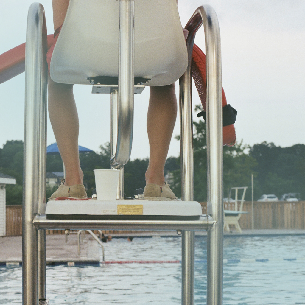 Ein Bademeister sitzt auf einem Hochstuhl und überwacht ein Schwimmbecken von oben, auf seinem Schoß liegt eine Rettungsweste.
