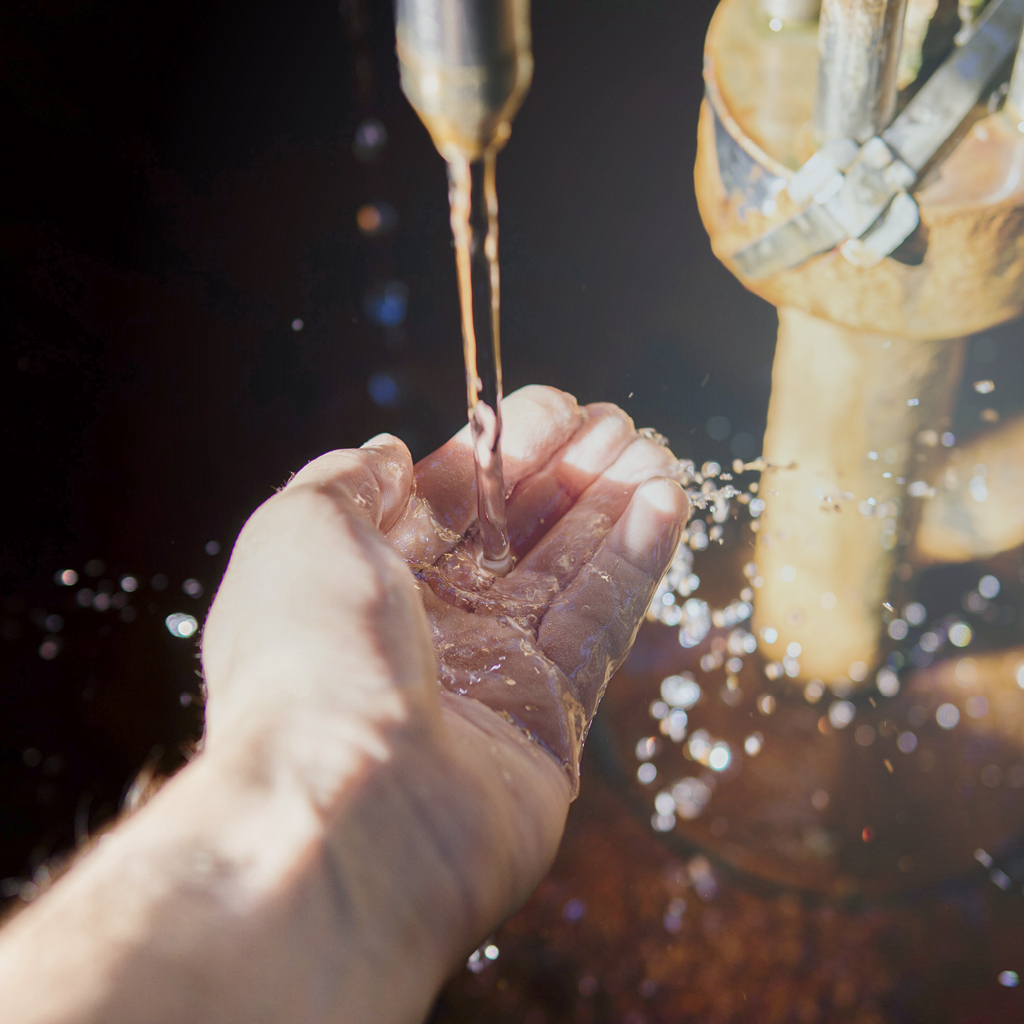 Eine linke Hand fängt Wasser mit der Handfläche auf und kontrolliert dieses, welches durch ein dahinterstehendes, vertikales  Rohr geflossen kommt.