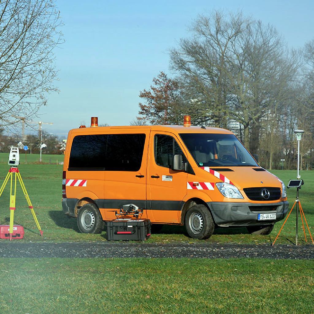 Ein oranges Vermessungsfahrzeug der Stadt Münster steht auf einer großflächigen Wiese, die ausgemessen werden soll.