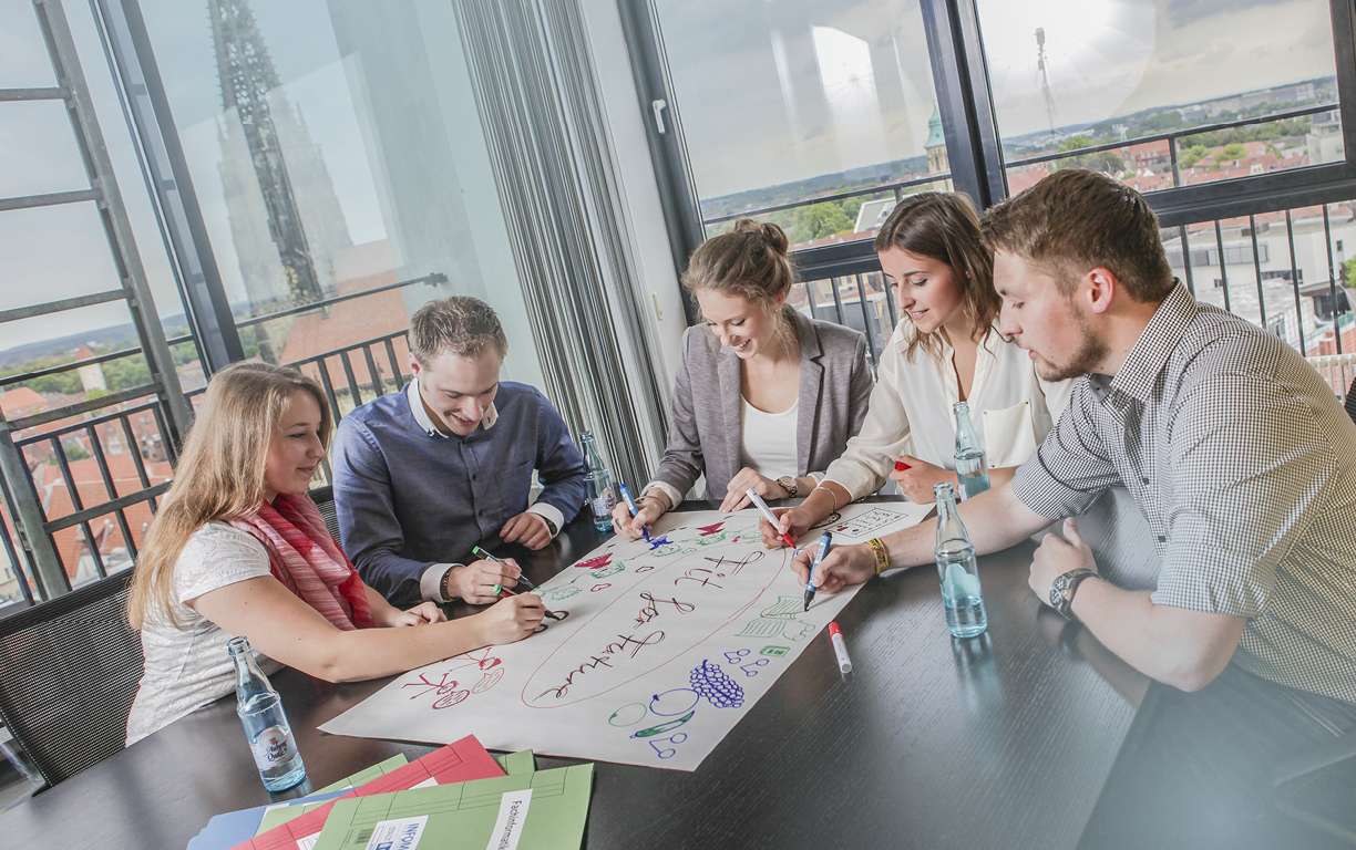 Eine Gruppe von fünf bunt gekleideten Männern und Frauen brainstormt gemeinsam an einem schwarzen Tisch in einem Workshop.