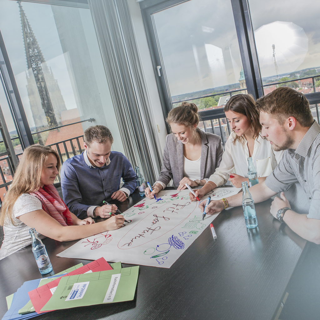 Eine Gruppe von fünf bunt gekleideten Männern und Frauen brainstormt gemeinsam an einem schwarzen Tisch in einem Workshop.