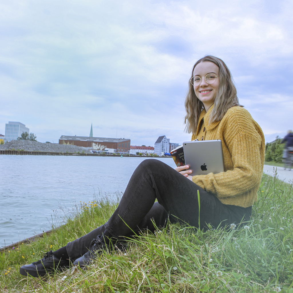 Eine Mitarbeiterin der Stadtbibliothek sitzt mit ihrem Arbeits-Tablet und einem Buch am Kanal und genießt ihre Mittagspause.