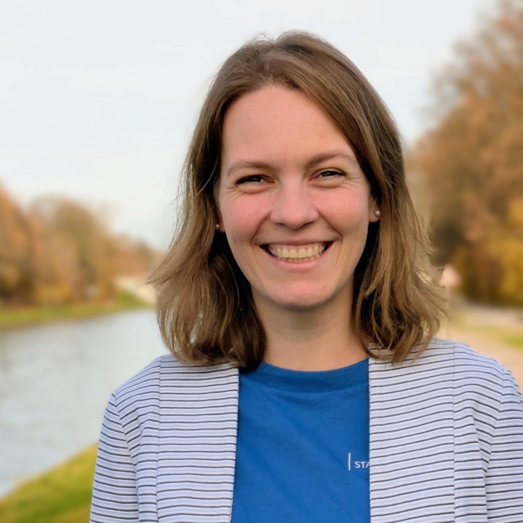 Eine Frau mit blauem Shirt steht am Kanal und lächelt in die Kamera.