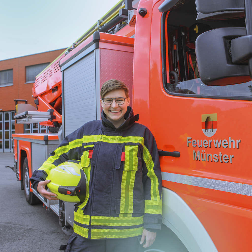 Eine Brandmeisterin steht stolz mit ihrer Ausrüstung vor einem orangen Drehleiter-Fahrzeug und lächelt in die Kamera.