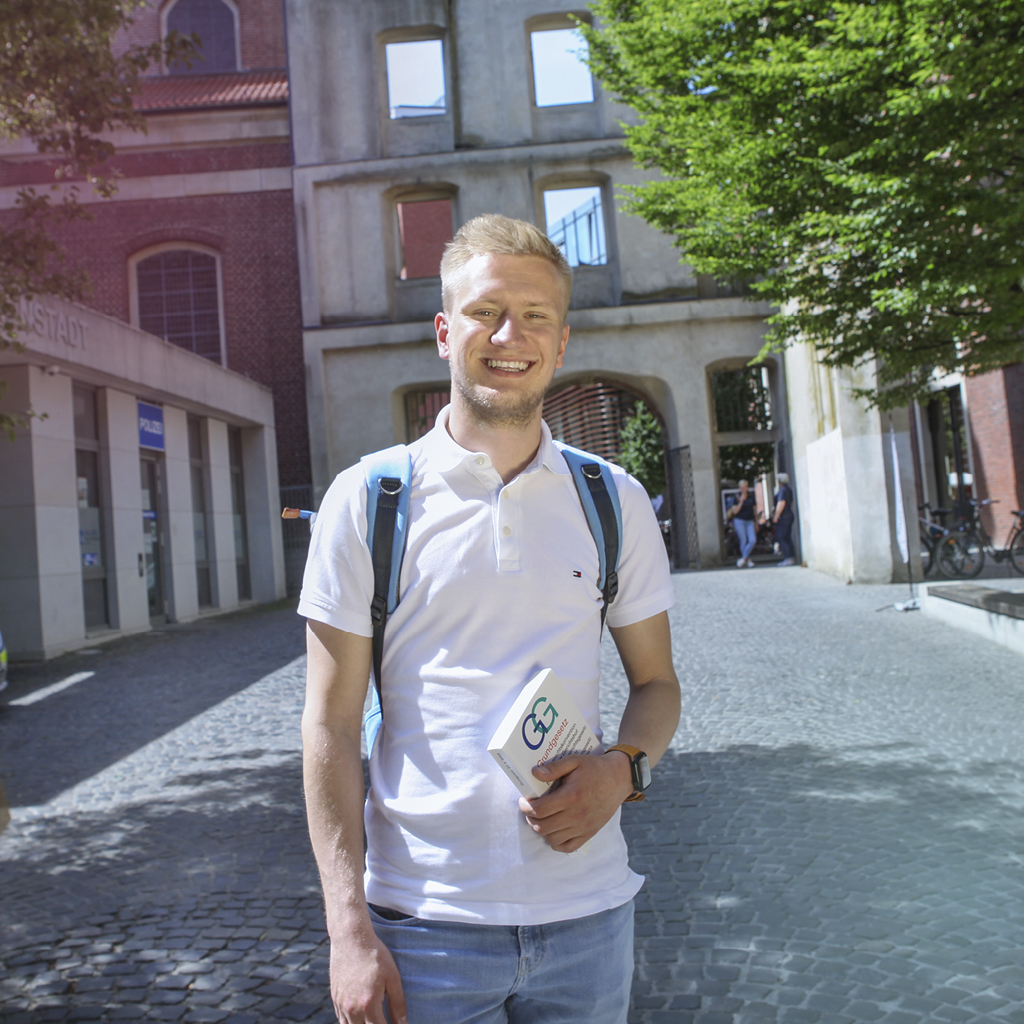 Vor einem architektonischen Denkmal in der Julius-Voos-Gasse steht ein angehender Verwaltungswirt und lacht in die Kamera.