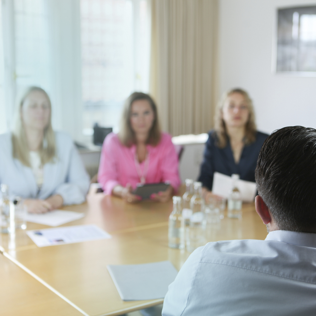 Ein Mann sitzt gegenüber von drei bunt gekleideten Frauen, die gemeinsam ein Bewerbungsgespräch führen.