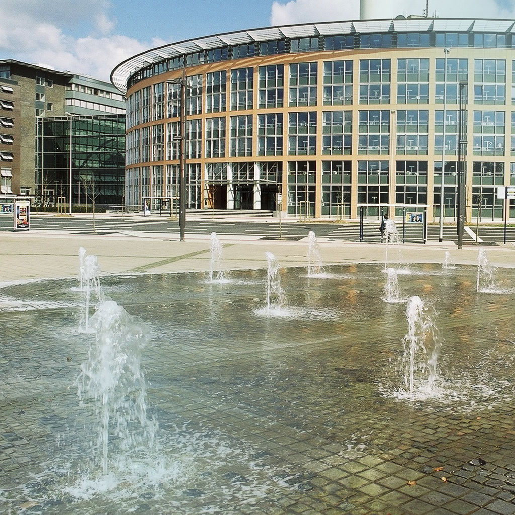 Ein Platz mit Wasserfontänen ist im Vordergrund zu sehen, dahinter befindet sich ein großen, rundes Gebäude der Stadt.