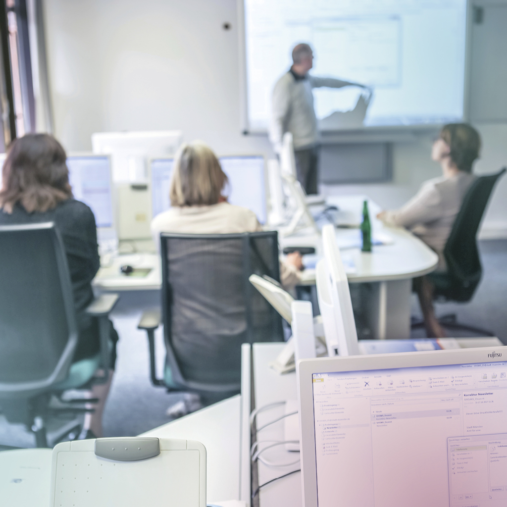 Mehrere Personen sitzen an eigenen Computern und arbeiten, im Hintergrund steht ein Mann an einer Leinwand und erklärt etwas.