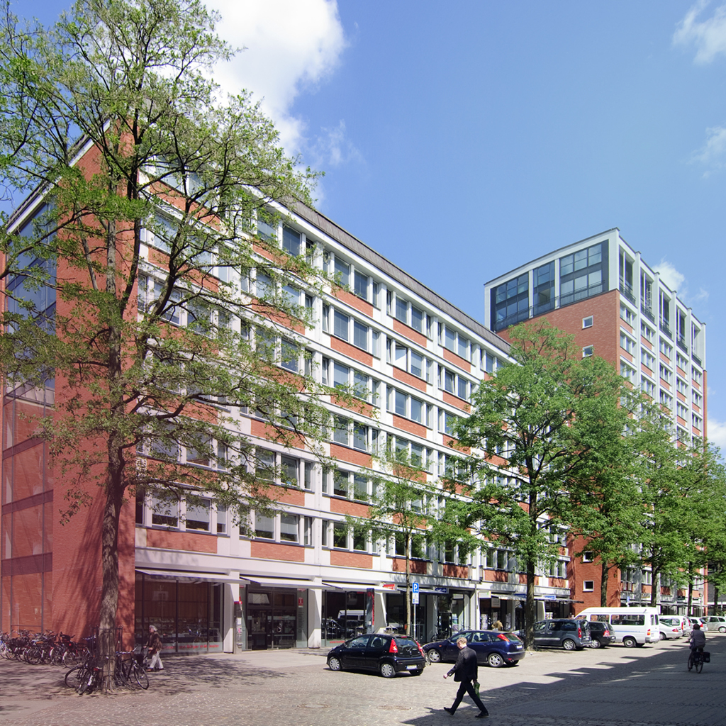 Hinter mehreren Bäumen ist ein gepflastertes Hochhaus zu sehen, in dem sich unter anderem die Kantine der Stadt Münster befindet.