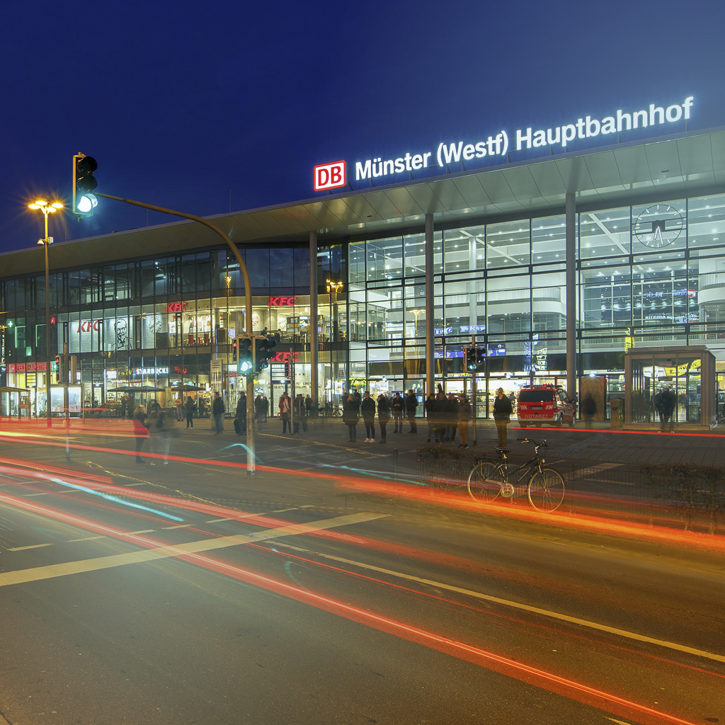 Ein Gebäude aus Glas wird in der Nacht von innen beleuchtet, vor dem Gebäude sieht man Lichtstreifen von schnell fahrenden Autos und Bussen.