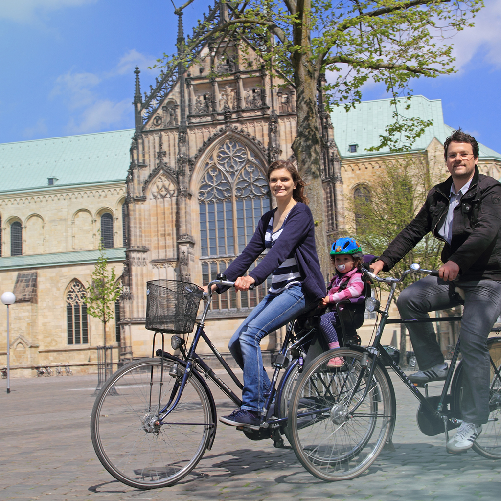 Eine Familie, bestehend aus zwei Eltern und einem kleinen Kind, fährt mit ihren Fahrrädern über den Domplatz von Münster.
