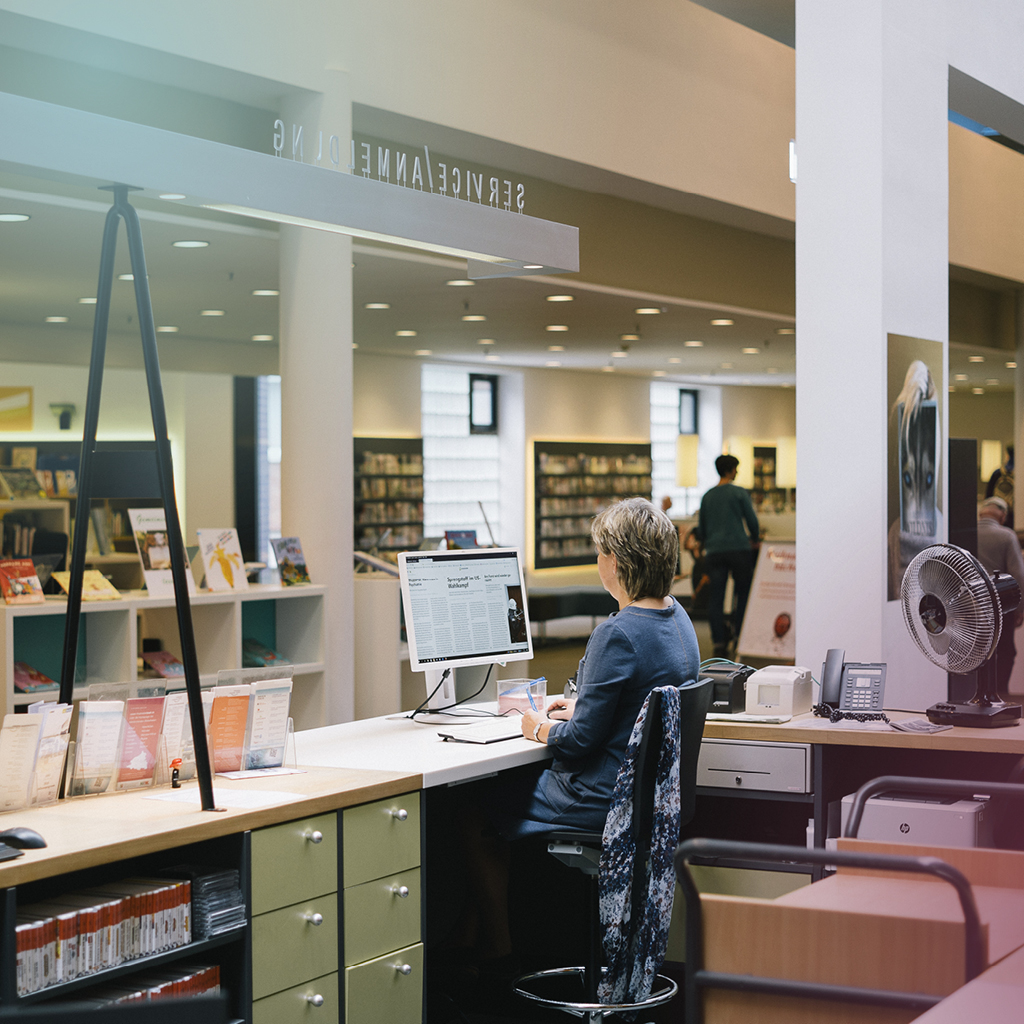 Eine Frau sitzt an einem Computer und im Hintergrund sieht man einige Menschen und viel Bücherregale