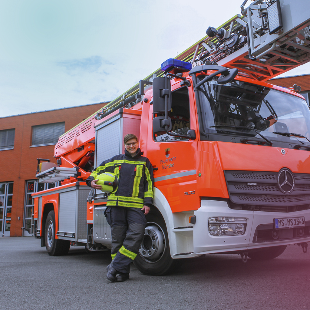 Eine Frau in Feuerwehruniform steht vor einem großen leuchtend orangen Leiterwagen vor dem Feuerwehrgebäude Münster