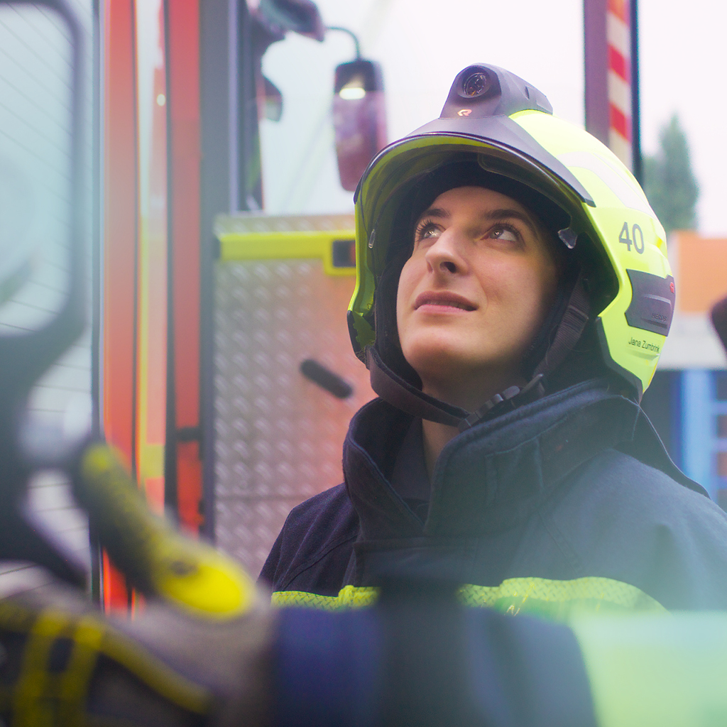 Eine Frau mit einem Feuerwehrhelm steht mit ihren Kollegen neben einem Einsatzwagen und schaut fragend nach oben