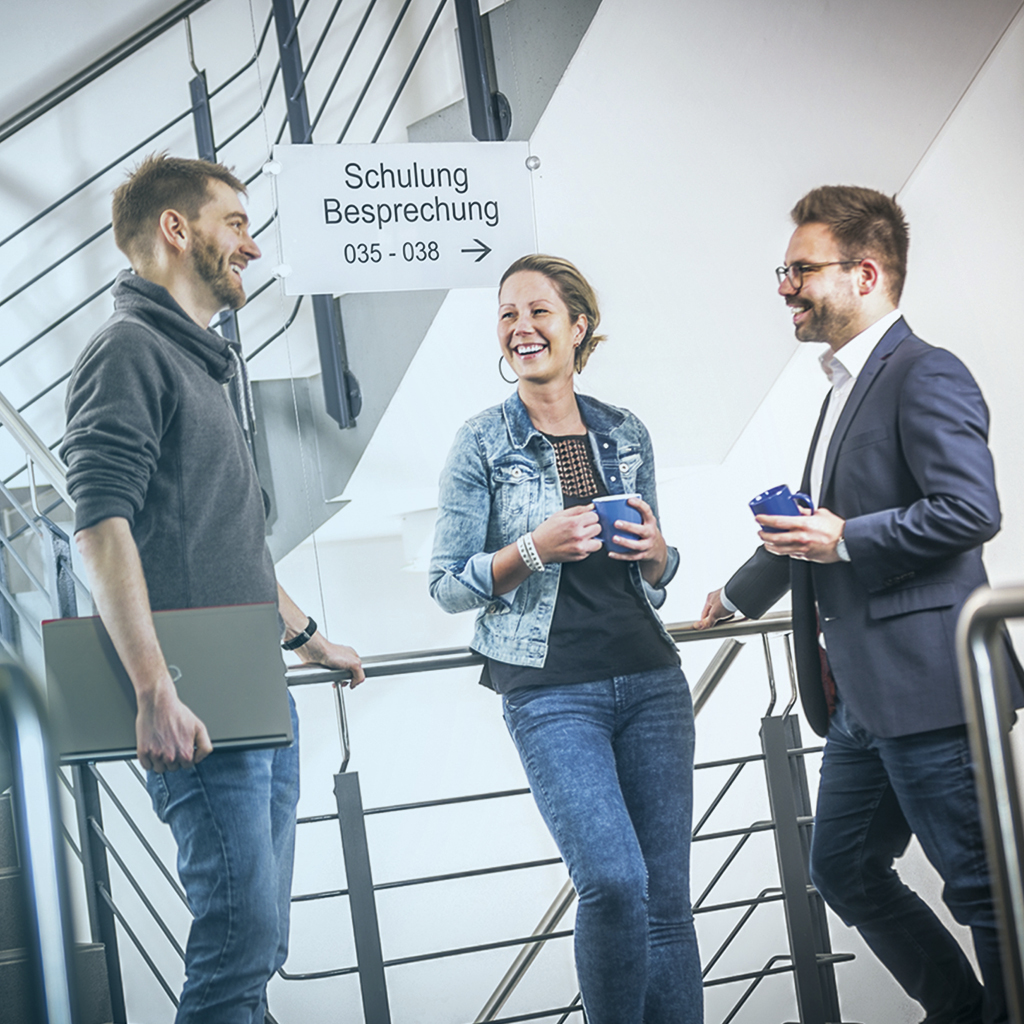 Drei Menschen treffen sich im Treppenhaus und reden miteinander undeiner hat ein Laptop unterm Arm