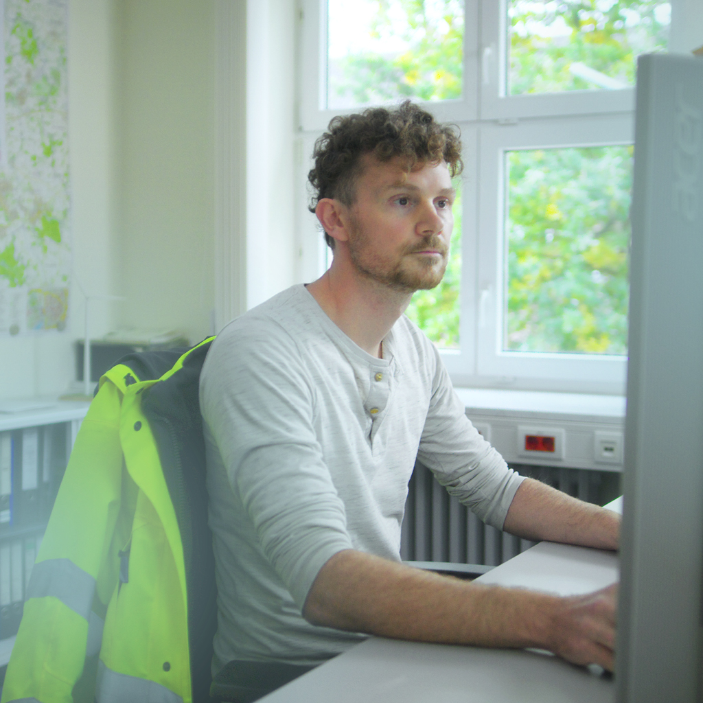 Ein Mann sitzt vor einem Computerbildschirm auf einem Stuhl, auf dem eine Sicherheitsjacke hängt und hinter ihm hängt eine Landkarte