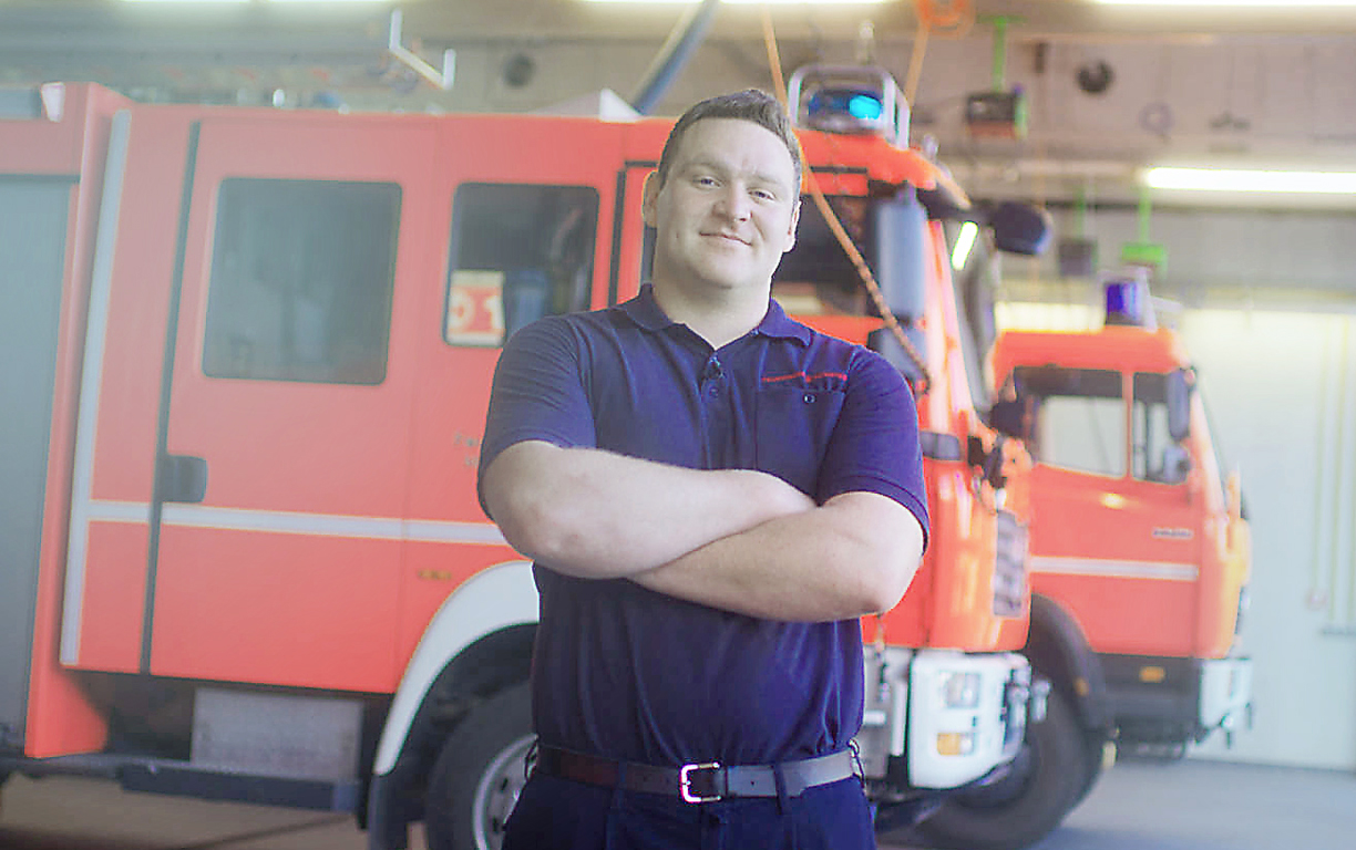 Ein Feuerwehrmann in blauer Kleidung steht mit verschränkten Armen vor zwei Einsatzfahrzeugen