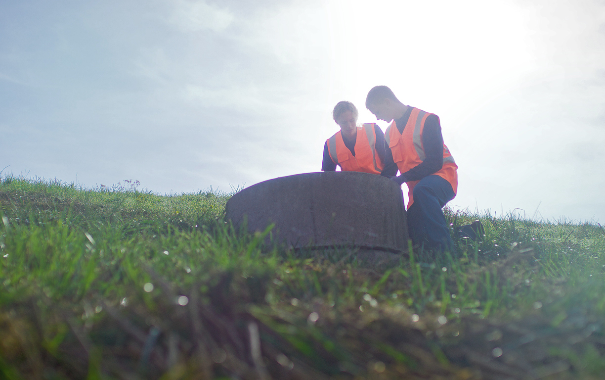 Eine Mitarbeiterin und ein Mitarbeiter kontrollieren einen runden Betonschacht auf einem grün bewachsenen Hügel