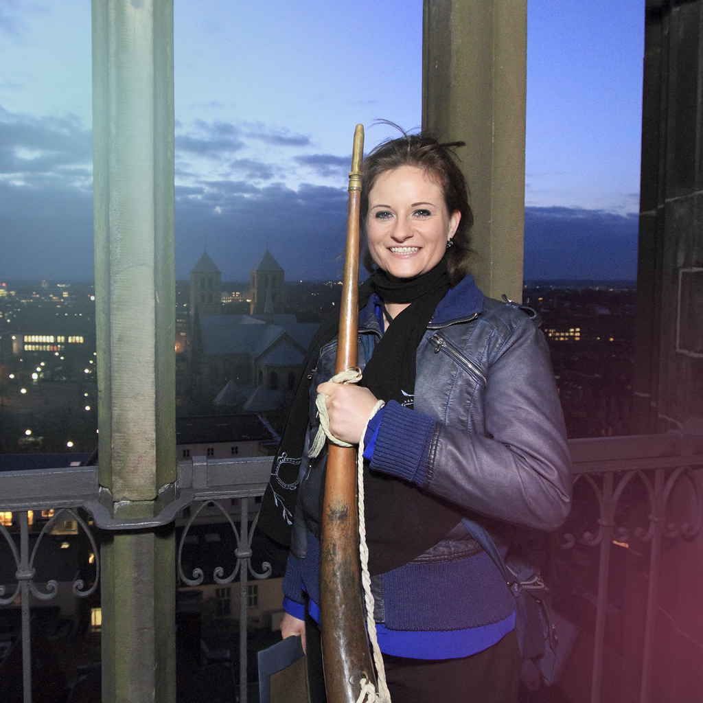 Eine Frau mit einem Blasinstrumen unterm Arm steht auf der Plattform eines Turms vor der nächtlichen Kulisse Münsters