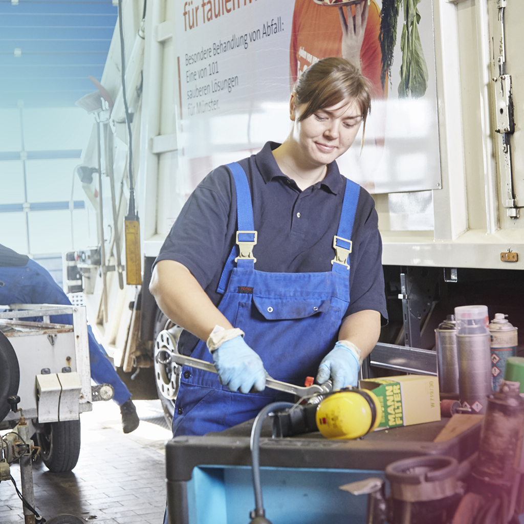 Eine KFZ-Mechatronikerin steht in einer Werkstatt, vor einem Abfallsammelfahrzeug, und repariert ein Teil davon.