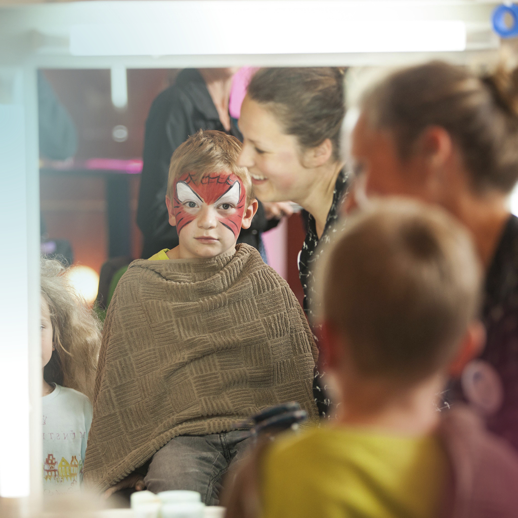 Eine Frau des Theaters schminkt einen Jungen mit roter Schminke im Gesicht.