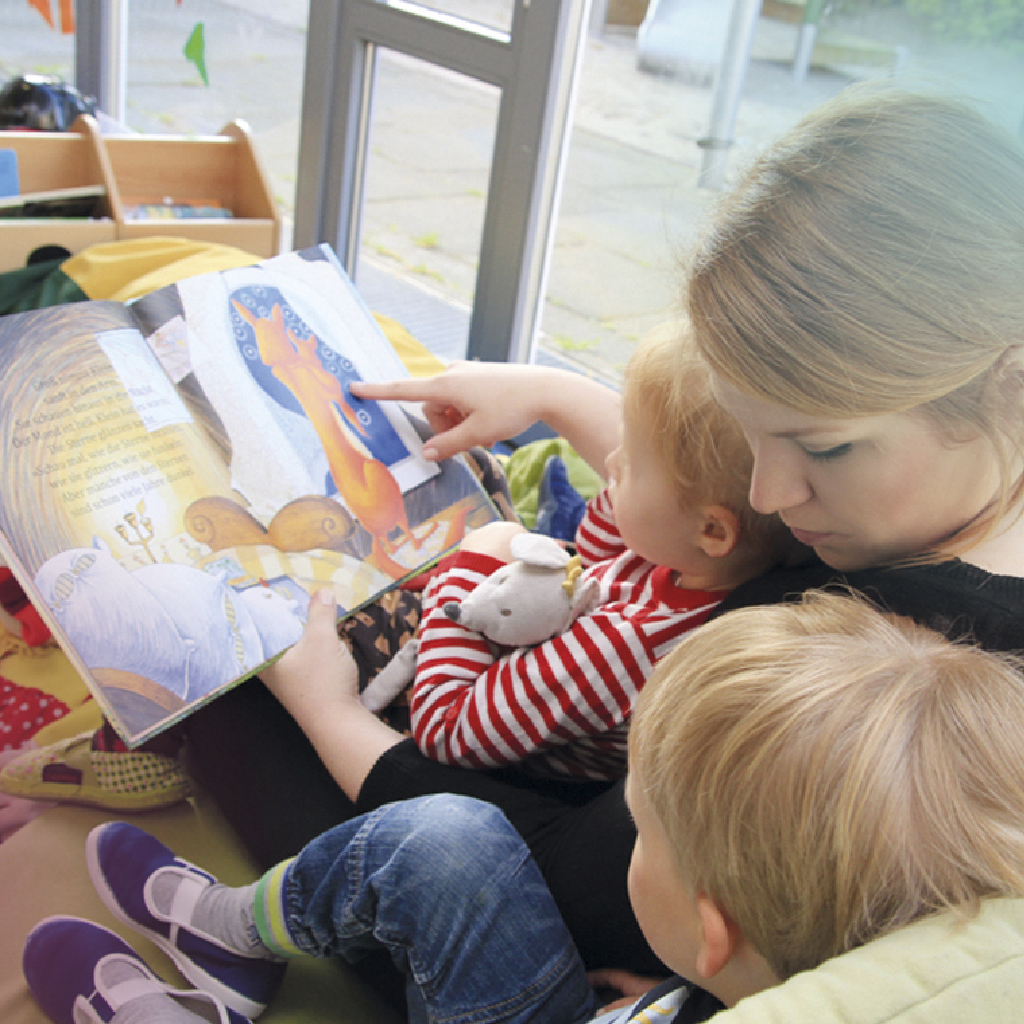 Eine Betreuerin einer Kindertagesstätte sitzt mit zwei Kindern auf dem Boden und liest ihnen etwas aus einem Buch vor.