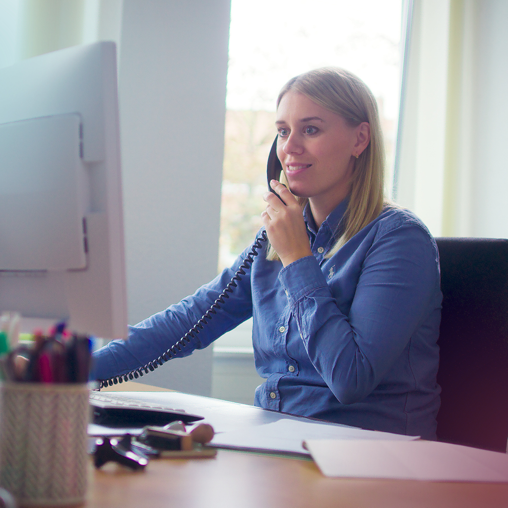 Eine junge Frau sitzt an einem Schreibtisch und telefoniert mit der einen Hand, während sie mit der anderen Hand am Computer arbeitet.