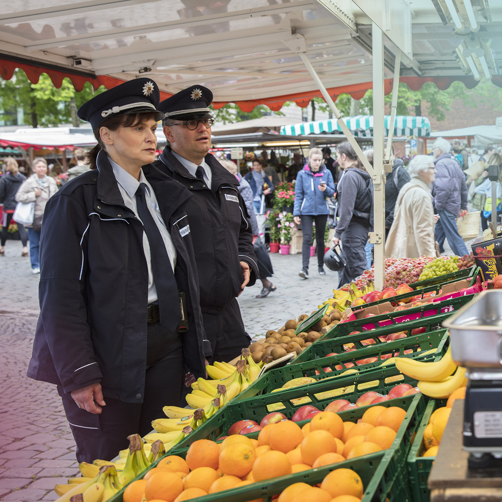 Zwei Kontrolleure des Ordnungsamtes stehen auf dem Markt in Münster und sprechen mit dem Besitzer eines Obststandes.