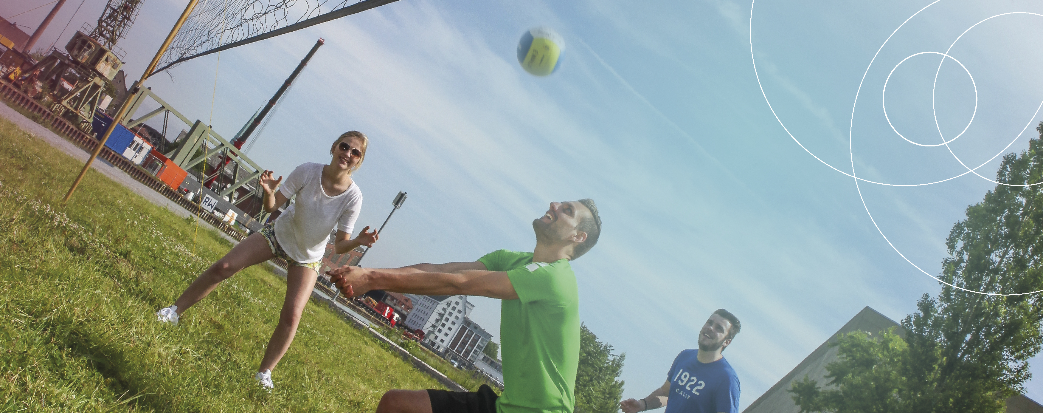 Zwei junge Männer und eine junge Frau zeigen sich sportlich gekleidet auf einer Wiese nahe dem Stadthafen und spielen gemeinsam Volleyball.