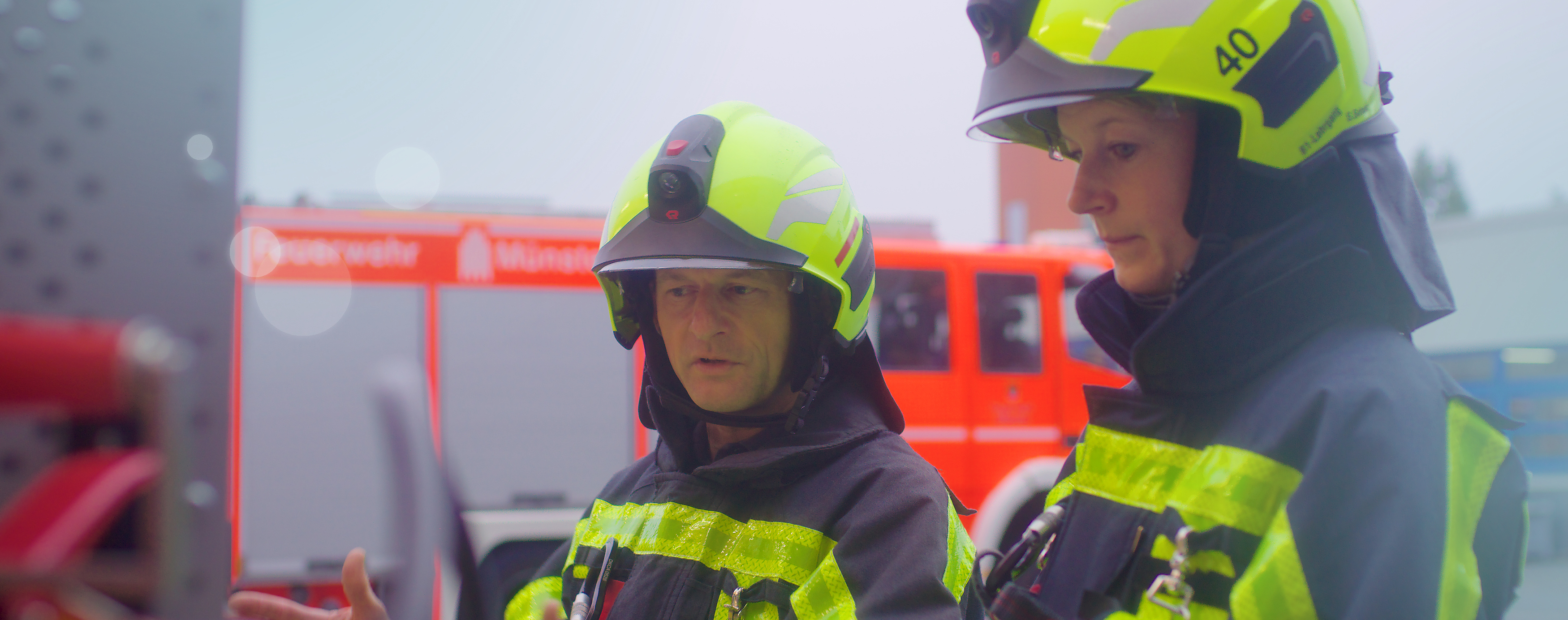 Zwei entsprechend gekleidete Feuerwehrleute stehen vor einem Fahrzeug und erklären einander etwas daran.