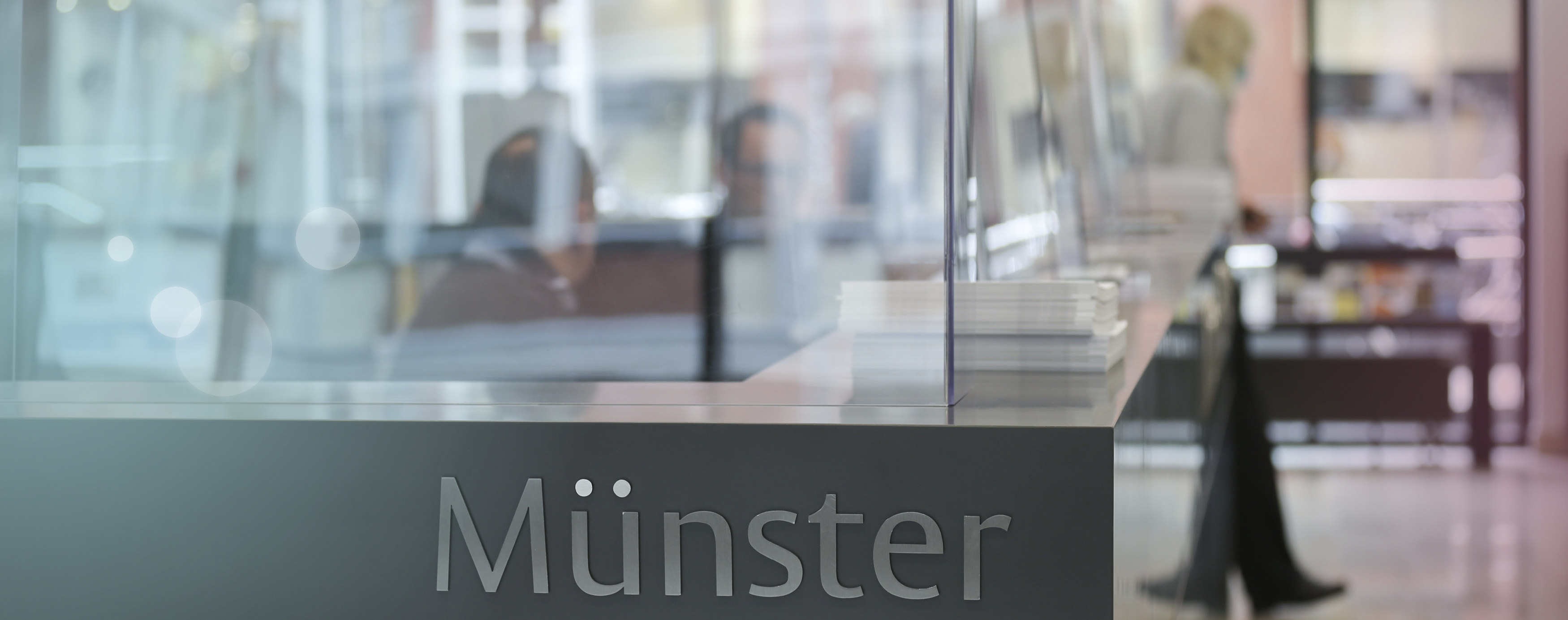 Eine graue Theke mit Glaswänden steht, mit dem Schriftzug "Münster" im Vordergrund, dahinter unscharf zu sehen sind Menschen die dort arbeiten und beraten.