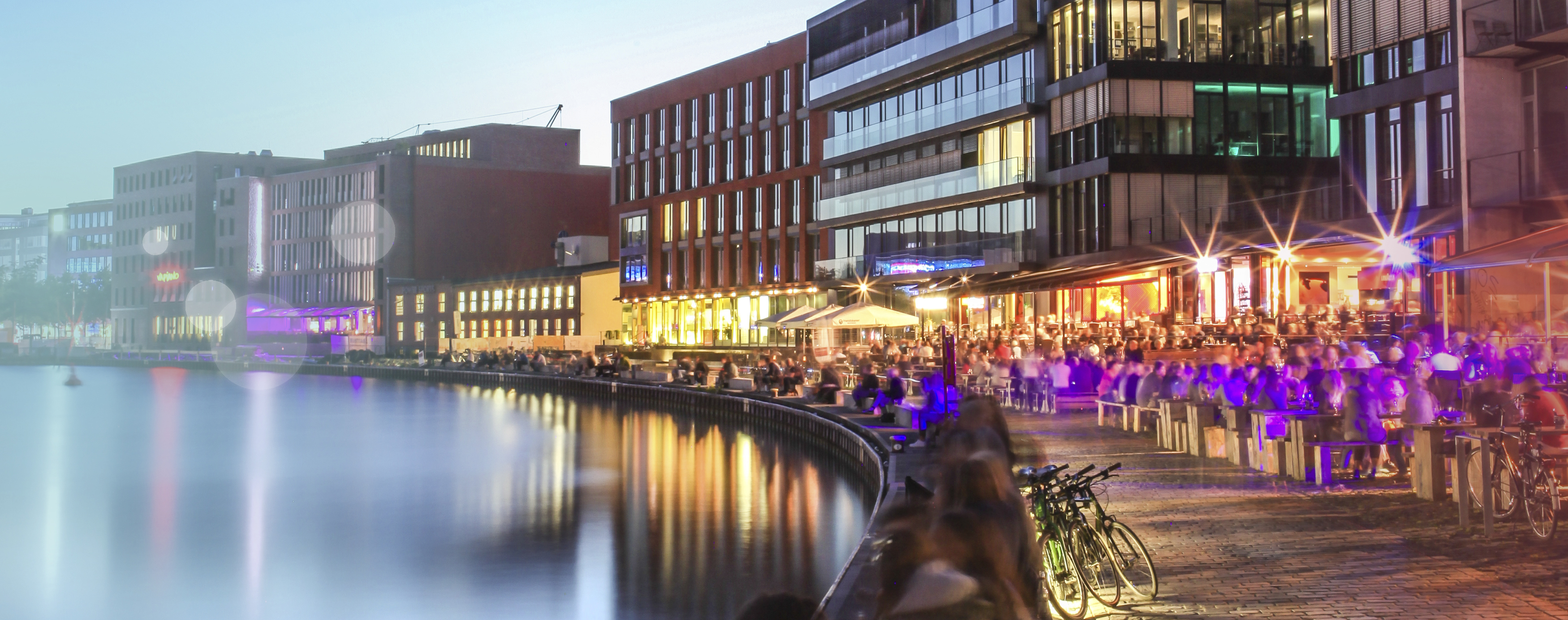 Der Hafen am Abend während des Sonnenuntergangs ist gefüllt mit Menschen, die in Lichtkegeln die Stimmung genießen.