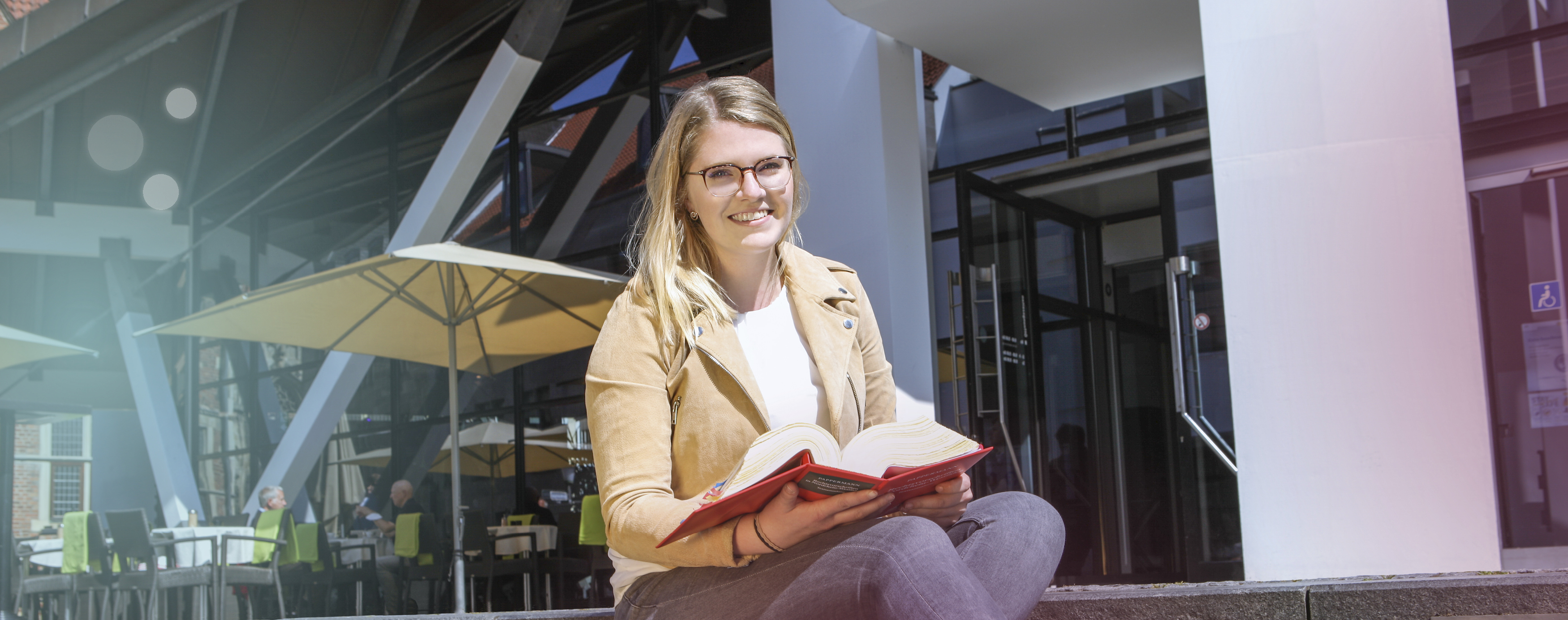 Eine blonde Frau sitzt auf der Treppe der Stadtbücherei Münster, schaut in die Kamera und blättert in einem Buch der Rechtsvorschriften.